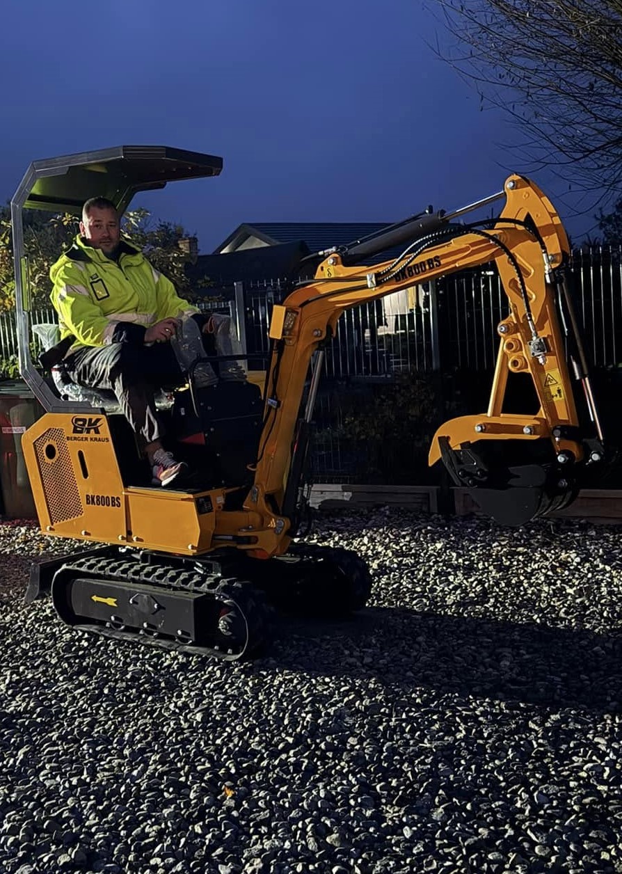 Zbliżenie na mężczyznę siedzącego w mini koparce na podwórku, patrzącego na kamerę.