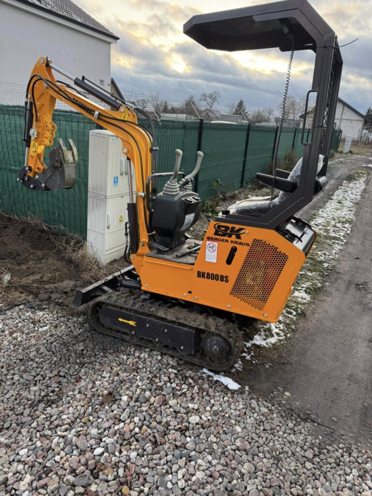 Mini koparka zaparkowana na poboczu drogi, gotowa do rozpoczęcia pracy.